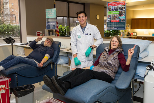 Two regular blood donors point their fingers up while sitting in the comfortable reclining chairs used during blood donations. A smiling technician is also shown in the photos.