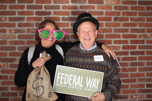 photo of a young woman with large, clown-like sunglasses on and a burlap bag with a big dollar sign on it in her hand, with her arm around the shoulders of an older man wearing a formal hat and patterned sweater who is holding a sign that says Federal Way Multi-Service Center. Both have big smiles.