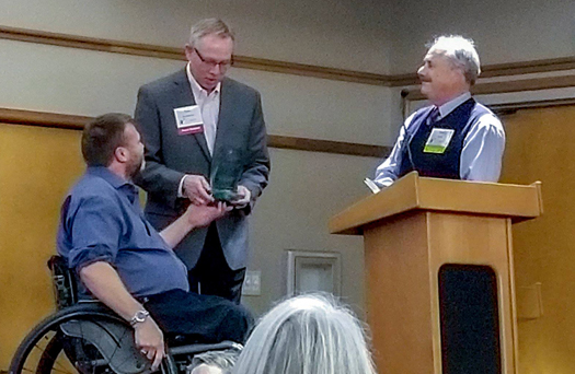 Photo shows Barry Long receiving an award. Barry is sitting in a wheelchair. The presenter is standing. Tom Minty is also standing behind a podium at the other side of the photo.