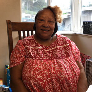 Photo of a siling African American woman with a bright red and white patterned dress and a yellow flower in her hair, smiling at the camera from her rocking chair.