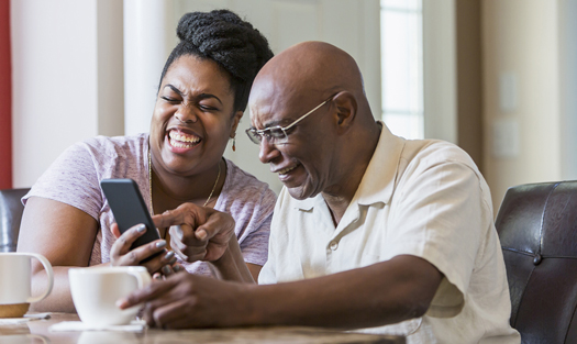 A senior African-American man in his 60s sitting at home with adult daughter, in her 30s at the dining room table, drinking coffee. The woman is showing her father something on her mobile phone and they are laughing.