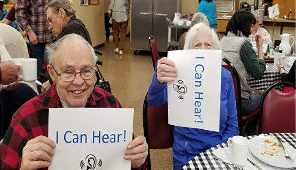two elderly people holding up I can hear! signs