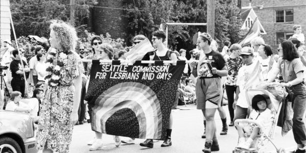An undated black and white photo show people marching for gay rights, holding a rainbow banner that reads "Seattle Commission for Lesbians and Gays."