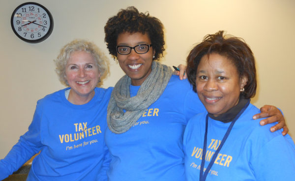 A white woman and two black woman stand together with their arms around each other's shoulders wearing matching blue shirts that say "tax volunteer."