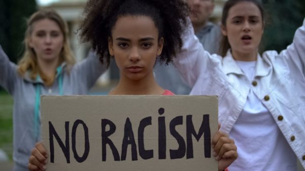young black woman holding up a cardboard sign with the words "no racism" written in black marker. Two young white women stand behind her with their arms raised.