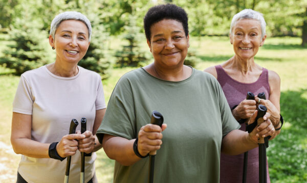 Waist up portrait of three sportive senior women smiling at camera holding walking poles outdoors
