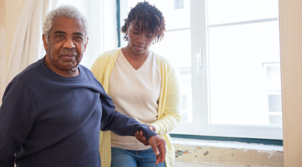The image depicts an older man and a younger woman in an indoor setting, possibly a room with a window. The man has gray hair and is wearing a dark blue sweater. He is looking at the camera with a calm expression. The woman, standing beside him, is wearing a cream or light yellow top and denim pants. She seems to be focused on holding or assisting the older man's arm, suggesting a caring or supportive gesture.
