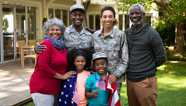Portrait of a young adult African American male soldier and a young adult mixed race female soldier with thier diverse multi-generation family in the garden outside their home, embracing and smiling to camera, a US flag draped over the shoulders of the kids