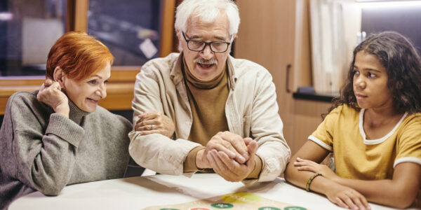 Mature man shaking dice in hands over board game while playing it with his wife and their cute interracial granddaughter