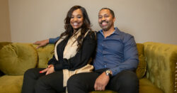 KD and David, a middle aged black couple, are pictured smiling and sitting on a green couch in their home.