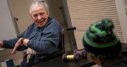 Kathleen is pictured sitting in a wheelchair and speaking with her caregiver, who is in the foreground wearing a beanie.