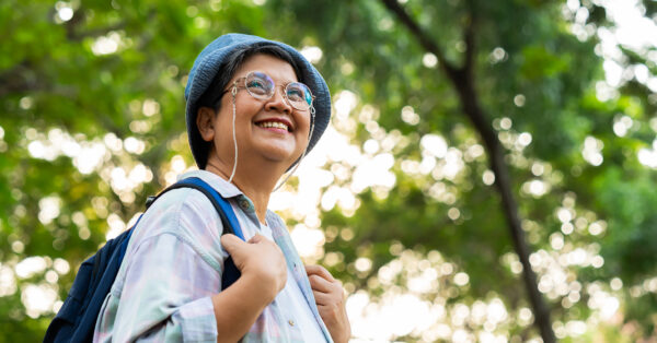 Portrait of Asian mature woman in a hat with backpack behind her back, an Asia active senior woman enjoying nature in autumn park. Standing on a trail in a forest outdoors. Enjoying active travel trip