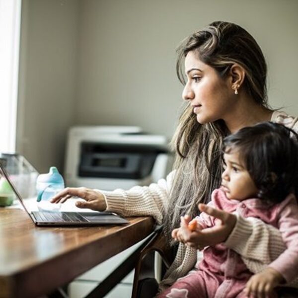 a mother holds her child in her lap while working on a laptop.
