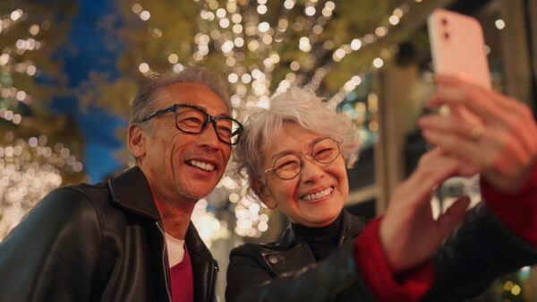 A senior couple capturing a joyful moment together as they take a selfie under a canopy of sparkling lights. Both are smiling, with the man striking a playful pose. The festive atmosphere of the illuminated evening street adds warmth and vibrancy to their cherished memory.