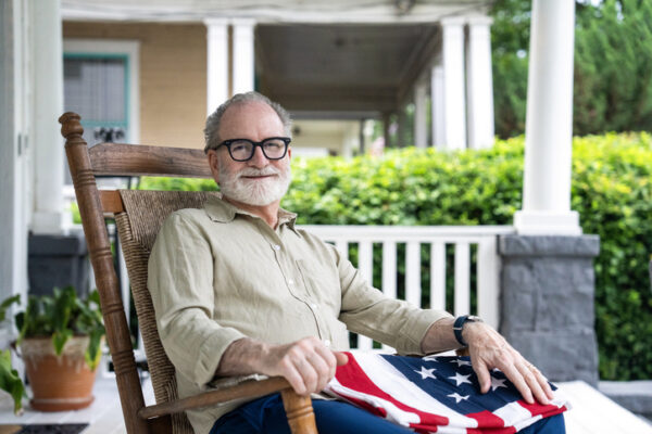 Side view of retired veteran looking at camera while sitting on his rocking chair on his porch with a flag on his lap.