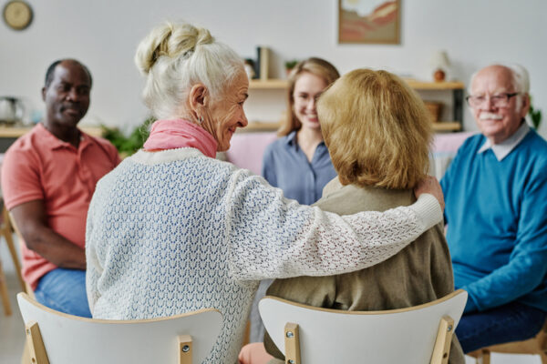 Rear view of senior woman supporting her friend while sitting at a support program meeting. With other older people.